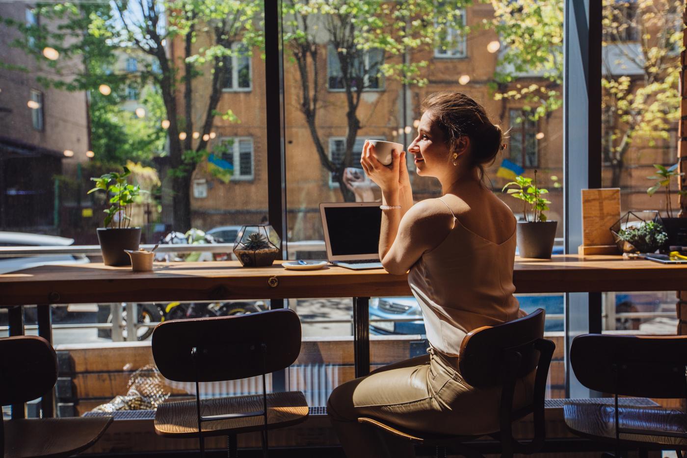 Frau entspannt mit Kaffee vor der Fensterfront in einem Cafe bei der Arbeit an ihrem Laptop