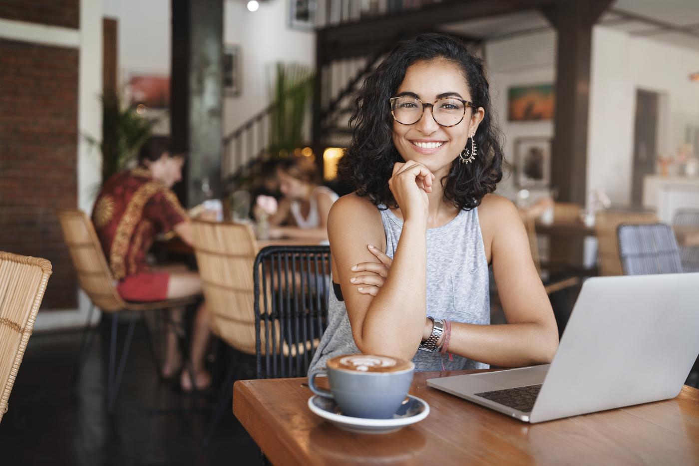 glückliche, entspannte Frau mit lockigem Kurzhaarschnitt und Brille, die am Laptop im Cafe arbeitet.