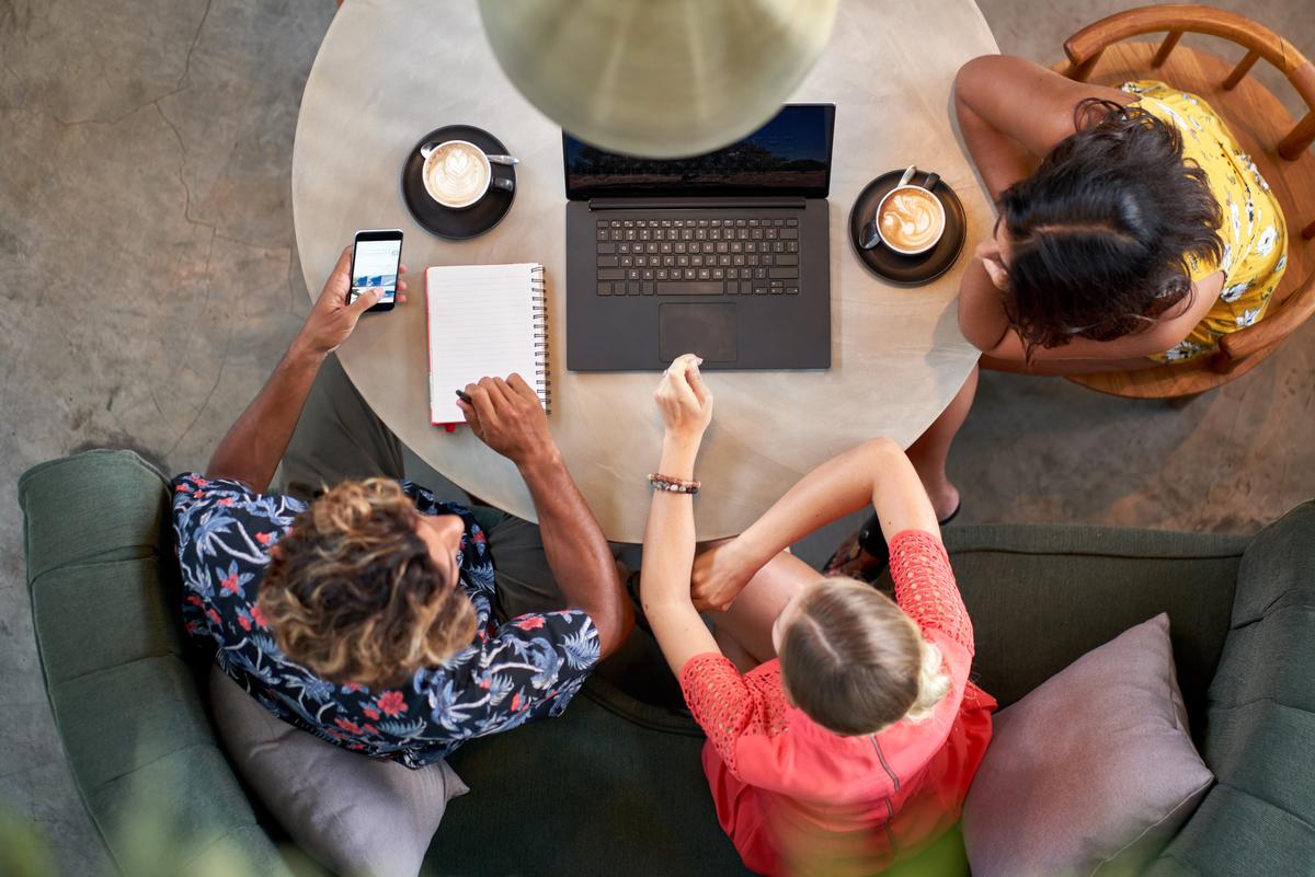 Drei Mitarbeitern, die in einem hellen Café, in dem fair gehandelter Kaffee serviert wird, bei einem Kaffee mit Laptop zusammenarbeiten